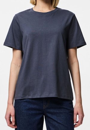 Persoon draagt een effen donkergrijs T-shirt met korte mouwen en donkerblauwe spijkerbroek, getoond van schouders tot heupen tegen een witte achtergrond.
