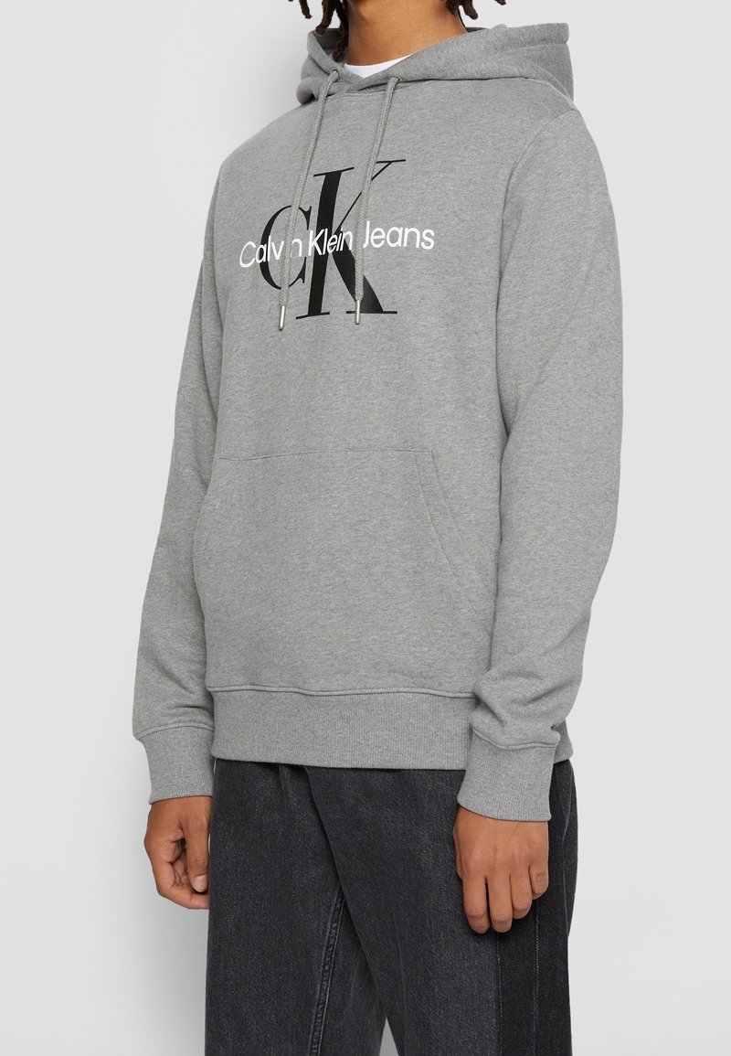 Szara bluza z kapturem, wykonana z miękkiego materiału, z kieszenią z przodu, sznurkami i dużym czarno-białym logo "Calvin Klein Jeans".