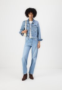 Giacca di jeans azzurro chiaro con collo e tasche frontali, abbinata a jeans azzurro chiaro a gamba dritta. Stivali con tacco marroni, mentre tiene una borsa beige.