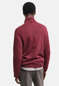 Maglione a maglia bordeaux con collo alto, colletto a costine e maniche lunghe, abbinato a una camicia a righe e pantaloni grigi.