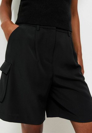 Personne portant un short cargo noir jusqu'aux genoux et un haut sans manches noir, avec une main dans la poche, sur un fond uni.