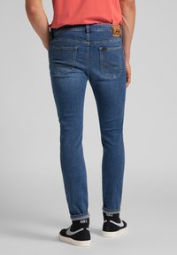 Jeans de denim azul de corte ajustado con un parche de cuero de la marca Lee, dos bolsillos traseros y un dobladillo enrollado. Combinados con zapatillas negras de Nike.