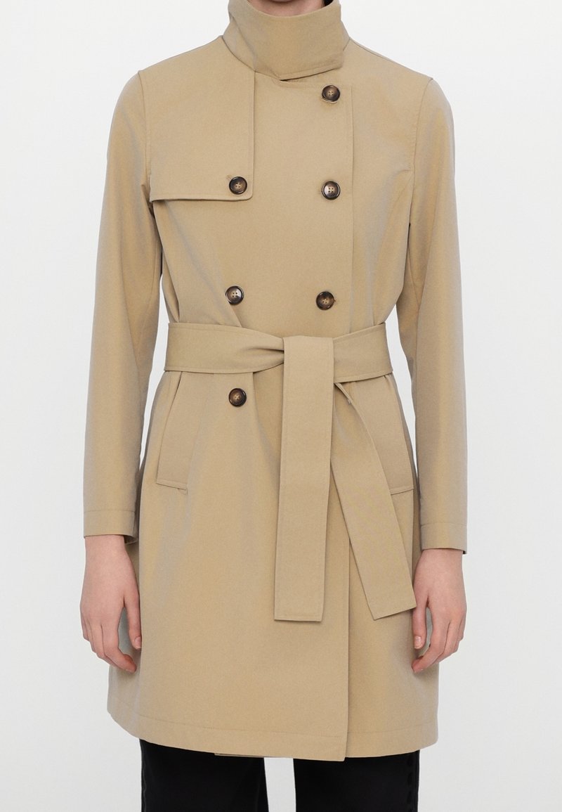 Trench-coat beige croisé avec des boutons foncés, col haut, ceinture nouée à la taille et poches latérales, porté sur un pantalon noir.