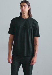 Tricou negru cu mâneci scurte și guler rotund, având un text subțire embosat. Asortat cu blugi închisi, expus pe un fundal deschis.