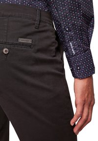 Zwarte katoenen broek met een achterzak met een klein label. Begeleid door een gestreept overhemd met lange mouwen in donkere tinten.