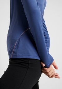 Top deportivo de manga larga en tejido azul oscuro, con paneles de malla texturizada, costuras en contraste y diseño ajustado. Combinado con leggings negros.