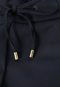 Tissu noir avec extrémités de cordon de serrage nouées, ornées d'embouts métalliques dorés gravés de logos.