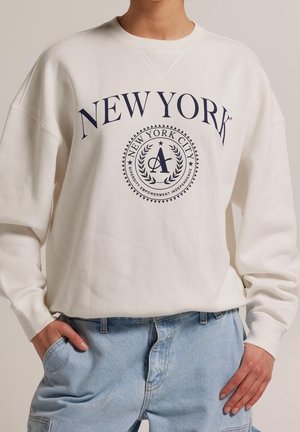 Person iført en hvid sweatshirt med "NEW YORK" og et rundt New York City-emblem, kombineret med lyseblå jeans, hænderne i lommerne.
