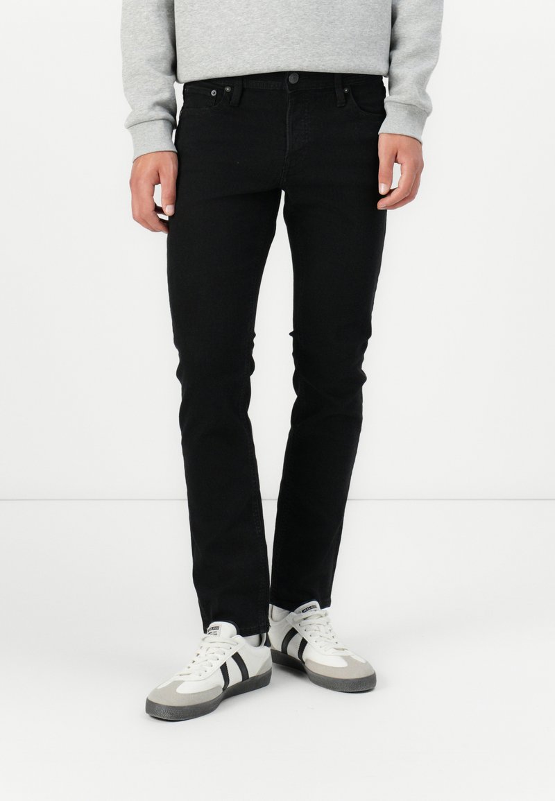 Jeans de mezclilla negra de corte ajustado, con cinco bolsillos y cierre de botón, combinados con zapatillas deportivas grises y blancas.