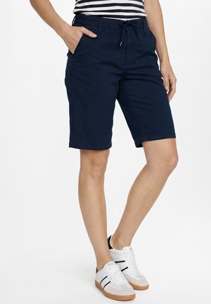 Person trägt marineblaue Shorts mit Kordelzug und Seitentaschen, ein gestreiftes Shirt und weiße Sneaker mit schwarzen Streifen.