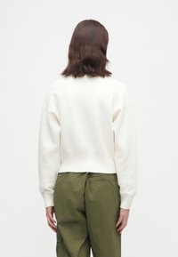 Osoba s hnědými vlasy po ramena, oblečená v bílé dlouhé svetr a olivově zelených kalhotách, viděná zezadu na jednobarevném pozadí.