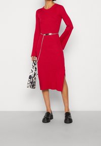 Robe tricotée rouge côtelée avec manches longues, fente latérale et ceinture chaîne argentée. Associée à une chaussure à plateforme noire et un sac à motif noir et blanc.