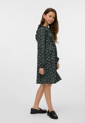 Sort blomstret kjole med en rynket halsudskæring, der har blå og grønne mønstre. Styleet med sorte loafers. Glat stof, knælangt design.