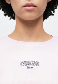 T-shirt rosa claro canelada com decote redondo e logótipo bordado "GUESS Jeans" em roxo escuro no peito. Textura lisa.