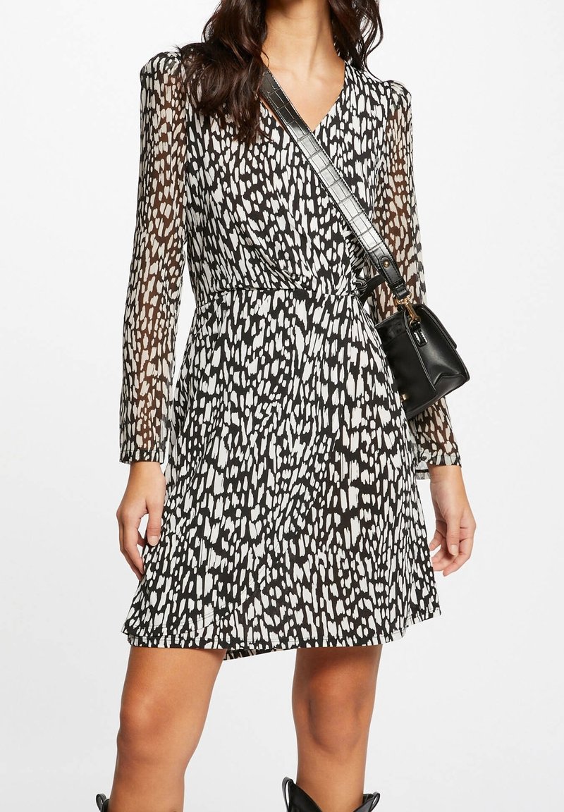 Femme portant une robe à manches longues noire et blanche à pois avec des manches transparentes, portant un sac bandoulière noir avec une sangle texturée.