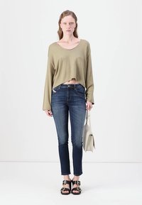 Suéter corto color verde oliva, jeans ajustados oscuros azules y sandalias negras con detalles de hebilla. Modelo sosteniendo un bolso de mano gris claro.