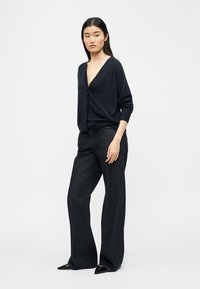 Vrouw met een zwart V-hals vest, zwarte broek met wijde pijpen en zwarte puntige schoenen, staand tegen een effen lichte achtergrond.