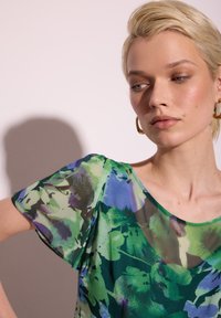 Kwiatowa bluzka w zielono-niebieskie wzory, wykonana z lekkiego materiału, z krótkimi rękawami i okrągłym dekoltem. Półprzezroczysta, o luźnym fasonie.