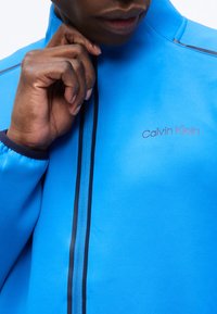 Άνδρας που φοράει φωτεινό μπλε μπουφάν Calvin Klein, με το χέρι κοντά στον λαιμό, κοντινή λήψη στο φερμουάρ και το λογότυπο στο στήθος.