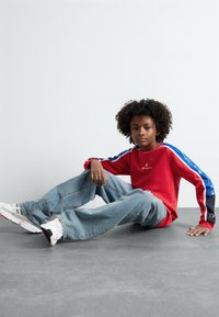 Camisa roja de manga larga con rayas de acento azules y negras, jeans de mezclilla azules y zapatillas blancas con detalles rojos y negros, sentado sobre una superficie gris.