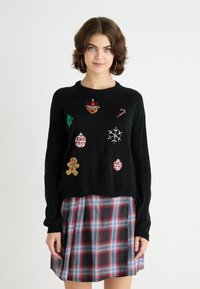 Pulover negru cu aplicații festive colorate, inclusiv ornamente, fulgi de zăpadă, turtă dulce și un brad de Crăciun. Guler și manșete striate.