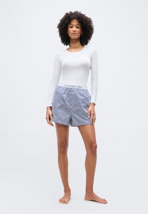 Femeie cu păr creț purtând un top alb cu mâneci lungi și pantaloni scurți Polo Ralph Lauren cu dungi alb-albastre, stând desculță pe un fundal simplu.