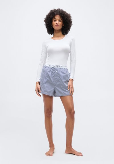 Femme aux cheveux bouclés portant un haut blanc à manches longues et un short Polo Ralph Lauren rayé bleu et blanc, debout pieds nus sur un fond uni.