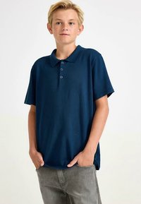 Marineblå poloshirt med korte ærmer, tre-knaps stolpelukning og krave, kombineret med lysegrå bukser.