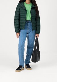 Zielona pikowana kurtka, zielona koszulka, jasnoniebieskie dżinsy, czarne trampki z białymi paskami i czarna torba na ramię na białym tle.