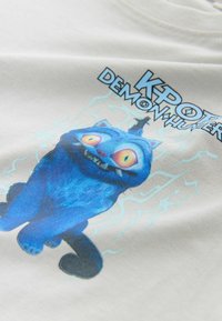 Weißes T-Shirt mit einer blauen Katzenfigur mit großen gelben Augen, die mit einem schelmischen Ausdruck sitzt, in elektrischem Blau umrandet.