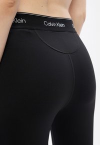 Čierne legíny s hladkým, elastickým materiálom, s širokým pásom označeným "Calvin Klein" a jemnými švovými detailmi.