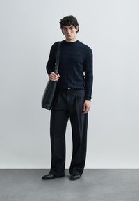 Črni pulover z rebrastim vzorcem, črne široke hlače in ujemajoči se črni natikači. Modelka nosi eleganten črn ramenski torbico. Minimalističen dizajn in čiste linije.