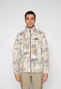 Mann iført en hel-zip, høy-hals fleecejakke med et dempet skogmønster og beige cargo-bukser, stående mot en ensfarget bakgrunn.