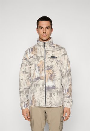 Homme portant une veste en polaire à zip intégral et col haut avec un motif forestier discret et un pantalon cargo beige, se tenant devant un fond uni.