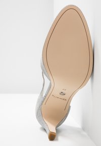 Semelle d'une chaussure à talon haut argentée avec un bout pointu, une semelle beige et un petit détail métallique près de la voûte plantaire.