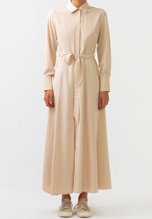 Frau trägt ein beiges, langärmeliges, geknöpftes Maxikleid mit einem gebundenen Taillengürtel und beigefarbenen Sneakern, vor einem schlichten weißen Hintergrund.