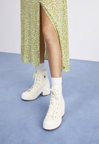 Bílé platformové sneakersy s vysokým střihovým designem a šněrováním, s bledým stříbrným kovovým ozdobením, nošené s bílými ponožkami, vedle zelených květovaných šatů. Modrá podlaha.