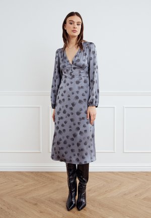 Femeie purtând o rochie midi gri cu mâneci lungi, cu modele florale negre, și cizme negre până la genunchi, cu vârf ascuțit, stând pe podea de lemn.