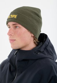 Olivgrüner Strickmütze mit gelbem Logo-Label, getragen mit einer marineblauen Jacke mit Reißverschluss und Kordelzugdetails. Nahaufnahme im Profil.