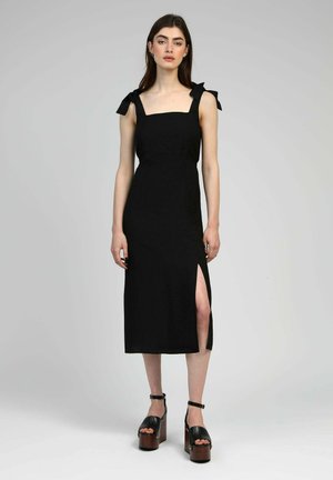 Femme debout portant une robe midi noire avec des bretelles nouées aux épaules et une fente haute sur la cuisse, accompagnée de talons compensés noirs.