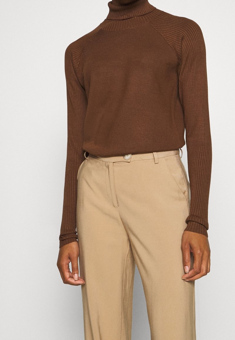 Pull à col roulé côtelé marron avec manches ajustées, associé à un pantalon beige doté d'une fermeture à boutons et de poches latérales.