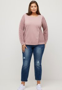 Vaaleanpunainen neulepaita, jossa on teksturoidut neulekuviot ja sivupaneelit; yhdistettynä keskisinisiin kuluneisiin farkkuihin ja valkoisiin lenkkareihin.