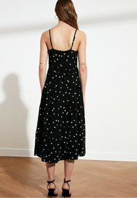 Czarna maxi sukienka w białe gwiazdy, na cienkich ramiączkach, z warstwowym wzorem i luźnym krojem. Noszona z czarnymi sandałami na kostkę.
