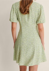 Robe florale verte avec des manches courtes bouffantes, un corsage ajusté et une jupe évasée. Le tissu présente un petit motif de fleurs dispersées.
