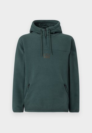 Hoodie en polaire vert foncé avec fermeture éclair sur le devant, deux poches latérales et un patch logo carré sur la poitrine. Texture douce.