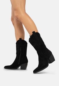 Las botas de cowboy de ante negro cuentan con una punta afilada, costuras western, cremallera lateral y un tacón cuadrado para mayor estabilidad y comodidad.