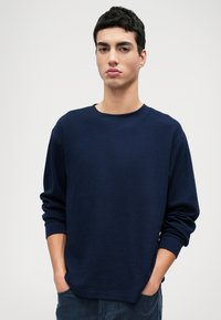 Námornícky modrý pulóver zo štruktúrovanej tkaniny, s okrúhlym výstrihom a dlhými rukávmi. Model stojí s rukami v vreckách.