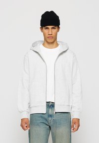 Ung mann iført svart lue, hvit zip-up hettejakke, hvit t-skjorte og falmede blå jeans, stående mot en nøytral bakgrunn.