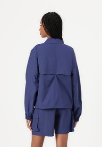 Marineblauwe lichte jas met een ontspannen pasvorm, elastische manchetten en een detail op de rug, gecombineerd met bijpassende shorts met cargozakken.