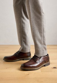 Zapatos de cuero marrón con cordones combinados con pantalones formales gris claro sobre un suelo de madera.
